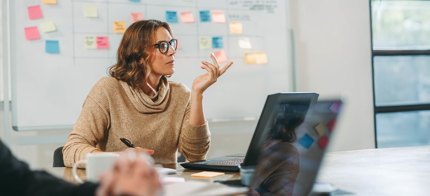 A woman sitting in a meeting room with other people and laptops having a brainstorming discussion. In the background is a whiteboard filled with sticky notes.